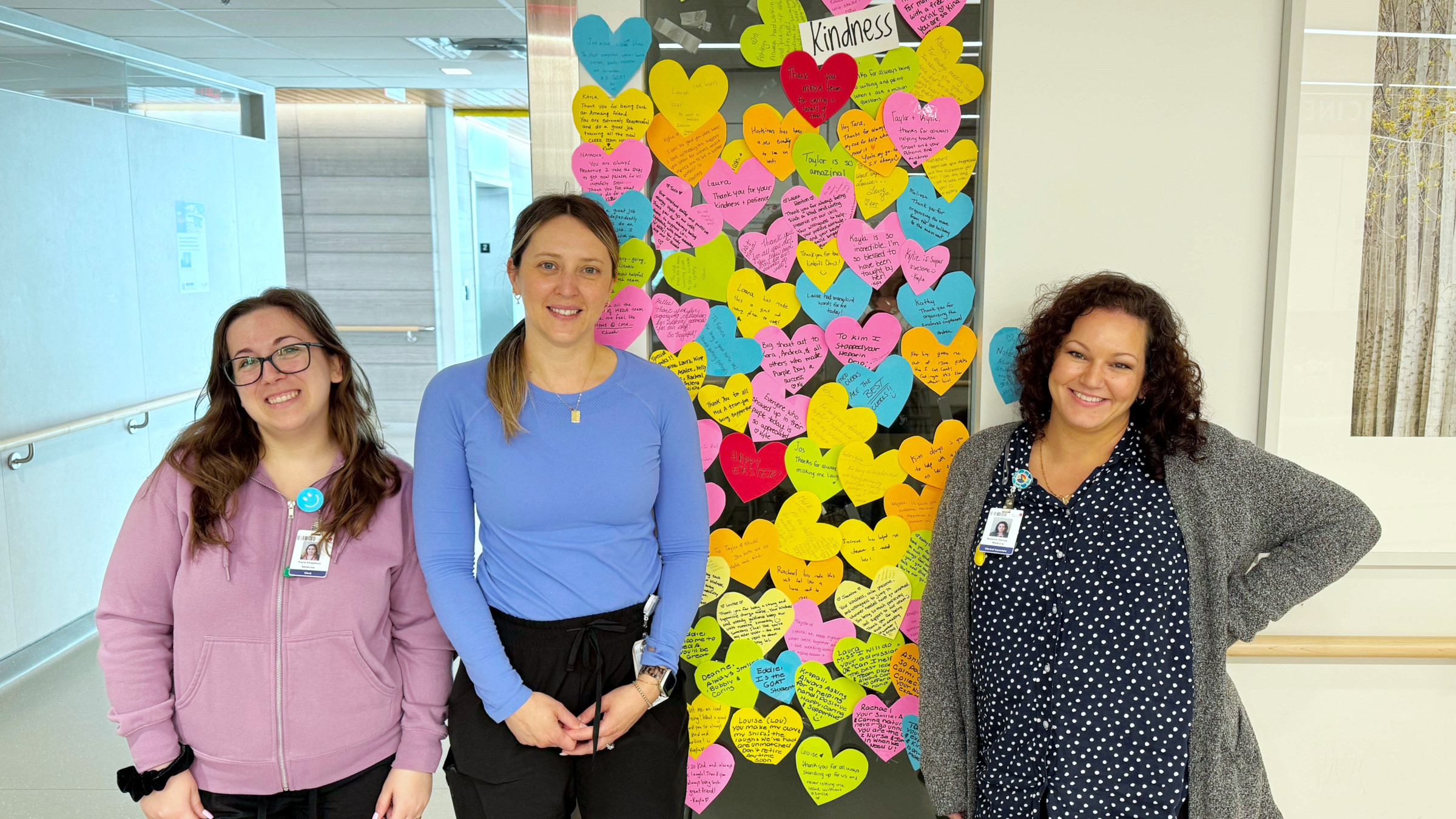 Three staff stand proudly in front of the kindness wall filled with post-its of messages to each other
