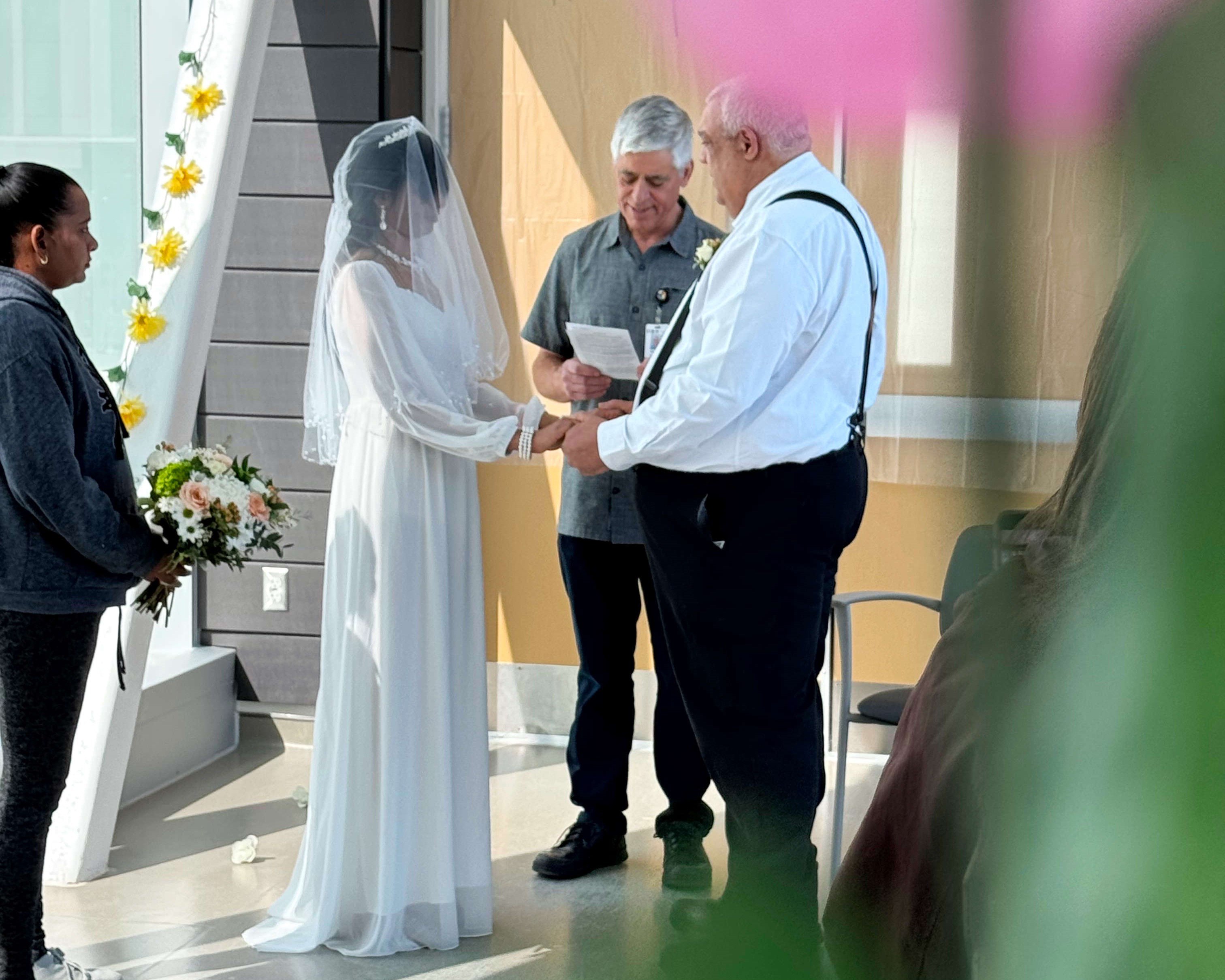 Alicia and Richard stand during their wedding ceremony, holding hands as the priest speaks